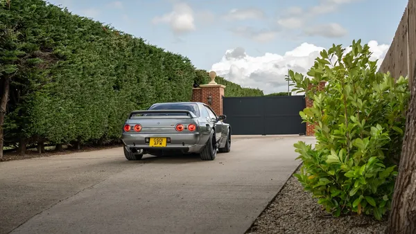 A grey sports car on a driveway facing a closed dark metal gate flanked by brick pillars and hedges