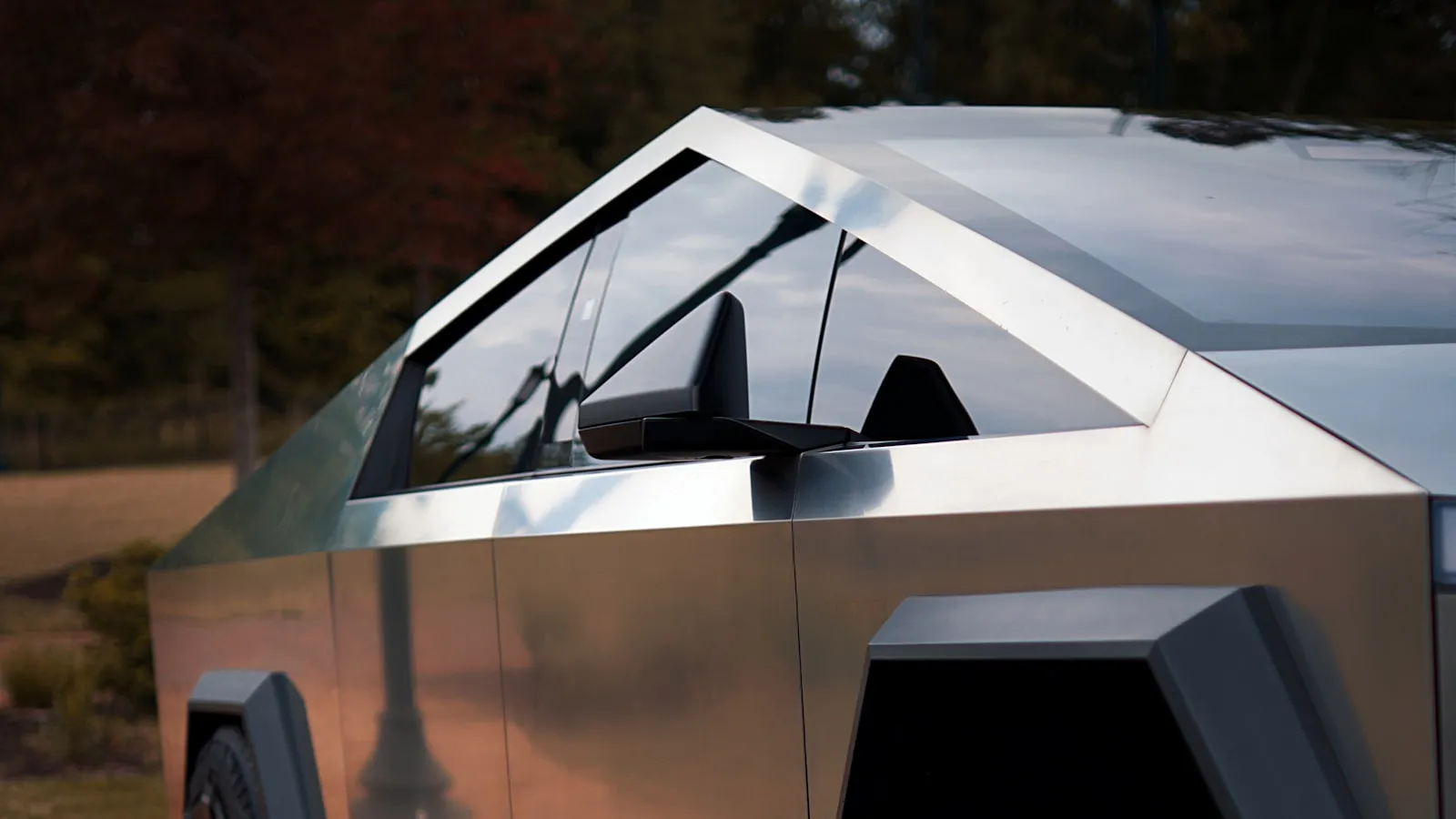 Side profile of a Tesla Cybertruck with stainless-steel exterior, foliage in the background