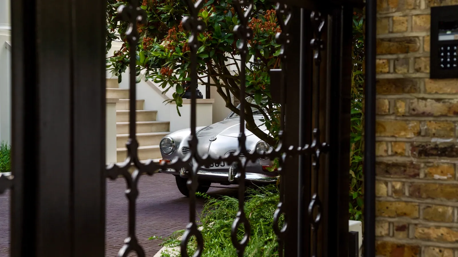 View through ornate wrought-iron driveway gate to a car on the driveway beyond, brick pillar with a keypad intercom panel at right