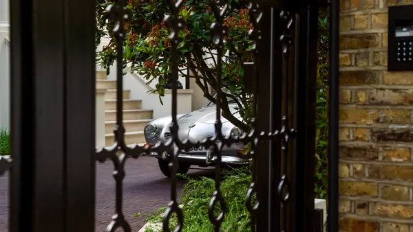 An ornate wrought-iron driveway gate open to a classic silver car on a driveway, a brick pillar with a keypad intercom at the right
