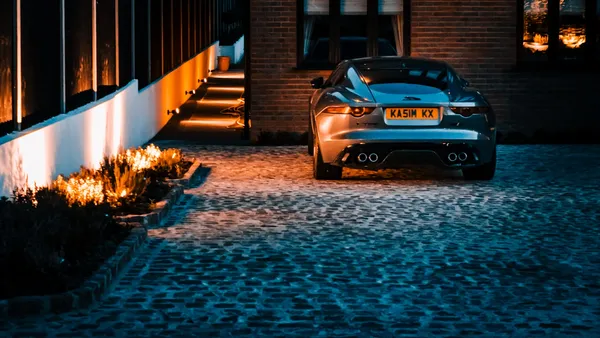 Silver sports car parked on a cobblestone driveway in front of a brick house, warm ground-level lighting at night