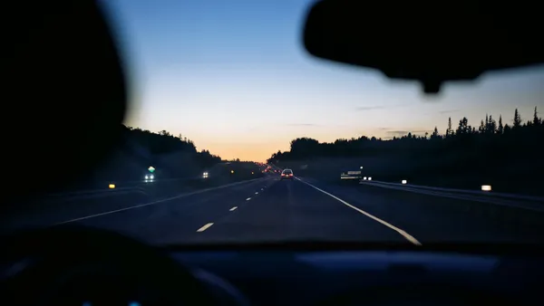 Windshield POV of a dusk highway with the rear-view mirror at the top and a tree-lined road stretching ahead