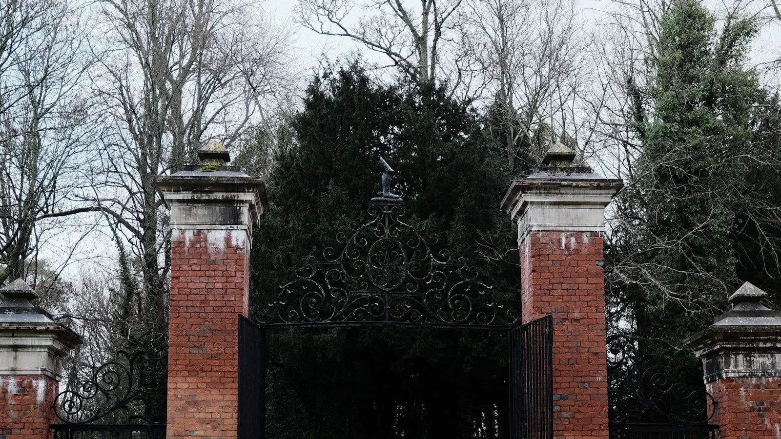 Ornate black wrought-iron gate between brick gateposts, bare winter trees in the background, overcast sky