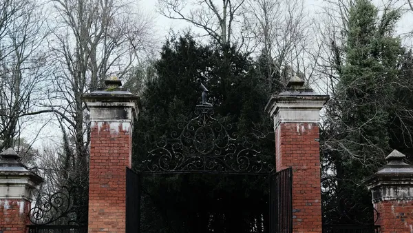 Ornate black wrought-iron gate between brick gateposts, bare winter trees in the background, overcast sky