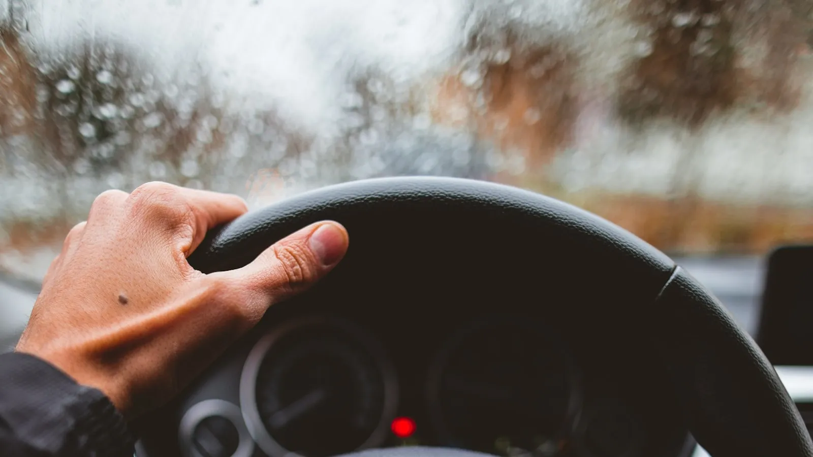 A hand gripping a steering wheel behind a rain-blurred windshield