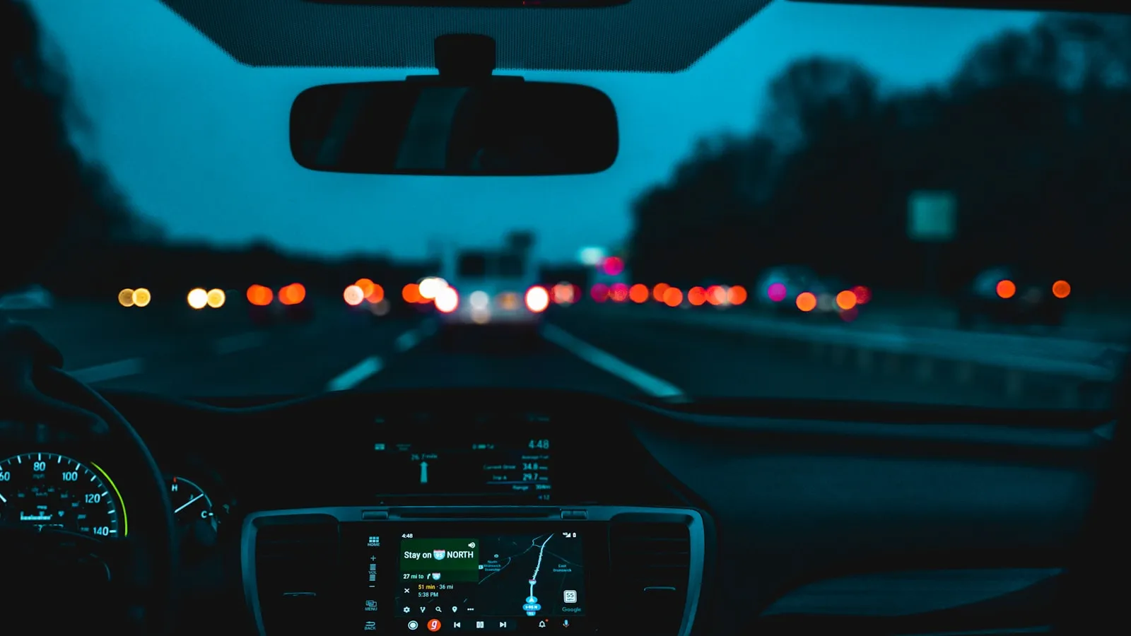 Dusk driver's-eye view inside a car showing the rear-view mirror and center navigation screen with blurred traffic lights ahead