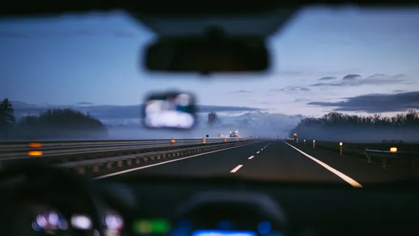 Dusk driver's-eye view through the windshield, a misty overcast road ahead with rear-view mirror visible at top and dash edge below