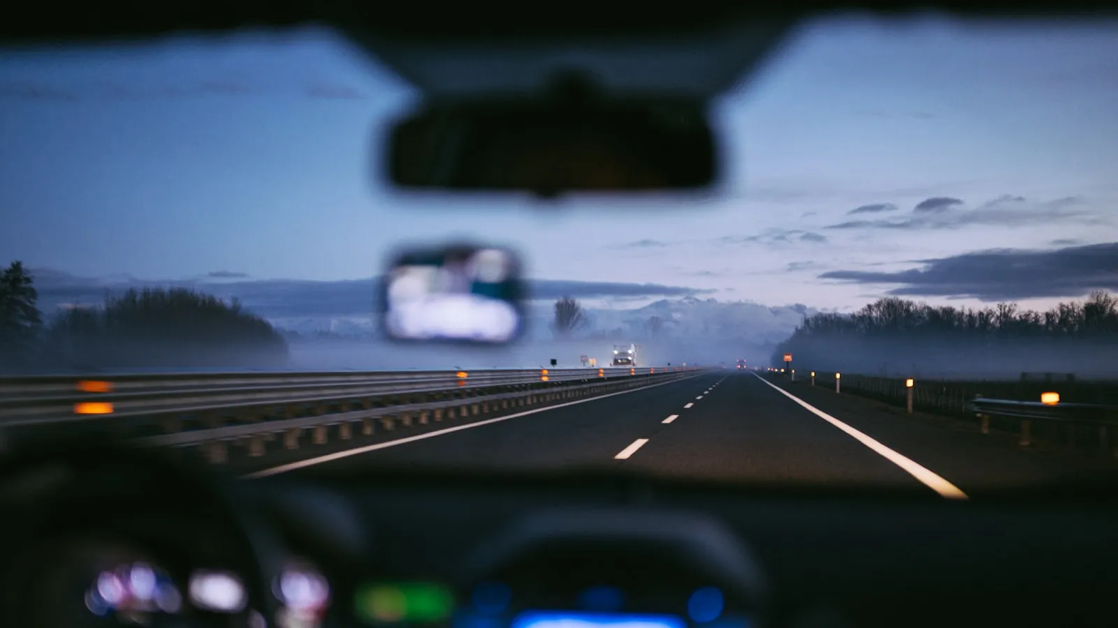 Driver's-eye view at dusk through a car windshield, rear-view mirror visible at top, blurred dashboard below, misty overcast road stretching ahead