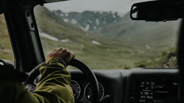 Overcast mountain drive from inside a car — hand on the steering wheel, instrument cluster showing gauges, center-console radio head unit visible