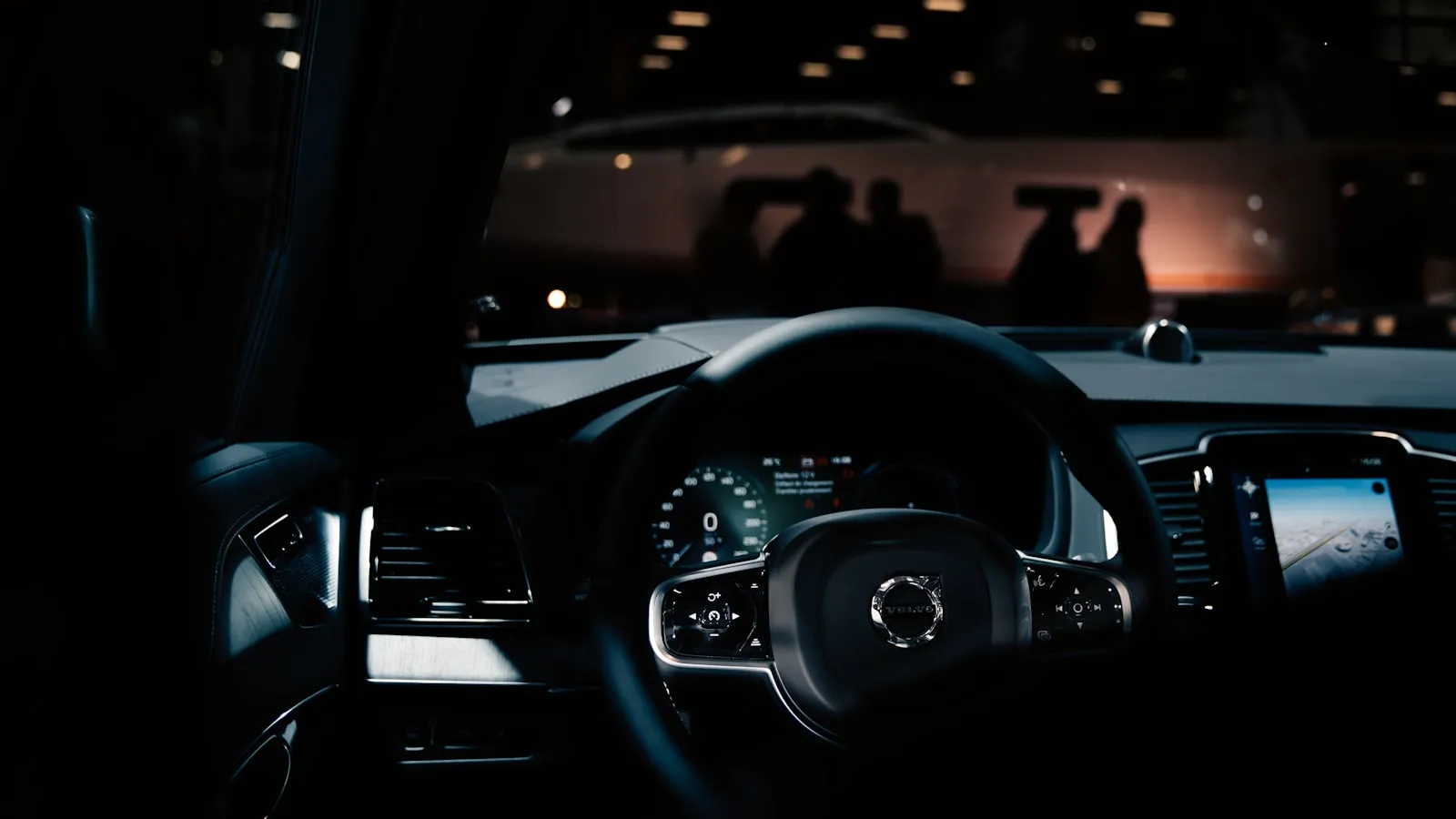 Dark Volvo steering wheel and instrument cluster glowing in a showroom, blurred lights behind