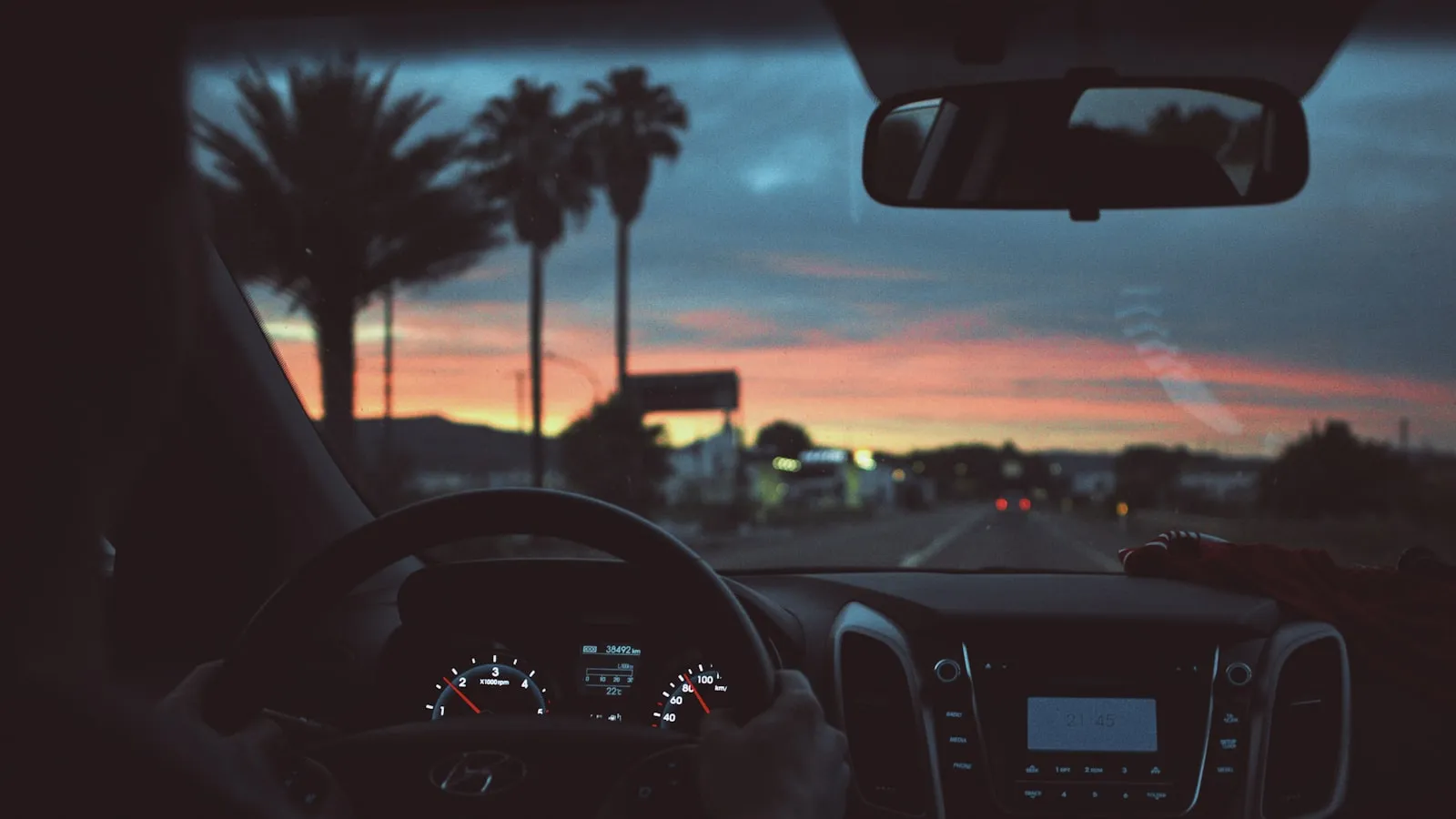 Driver POV with instrument cluster and rear-view mirror, palm trees and a pink sunset through the windshield