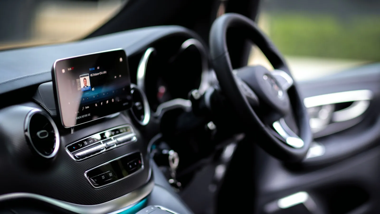 Car dashboard and steering wheel with a central infotainment touchscreen, greenery blurred through the windows
