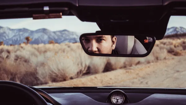 Rear-view mirror reflecting a driver's eyes, a small clock on the dashboard, desert landscape visible through the windshield