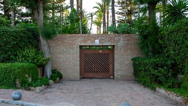 A wooden driveway gate set in a brick wall, framed by palm trees and lush tropical planting