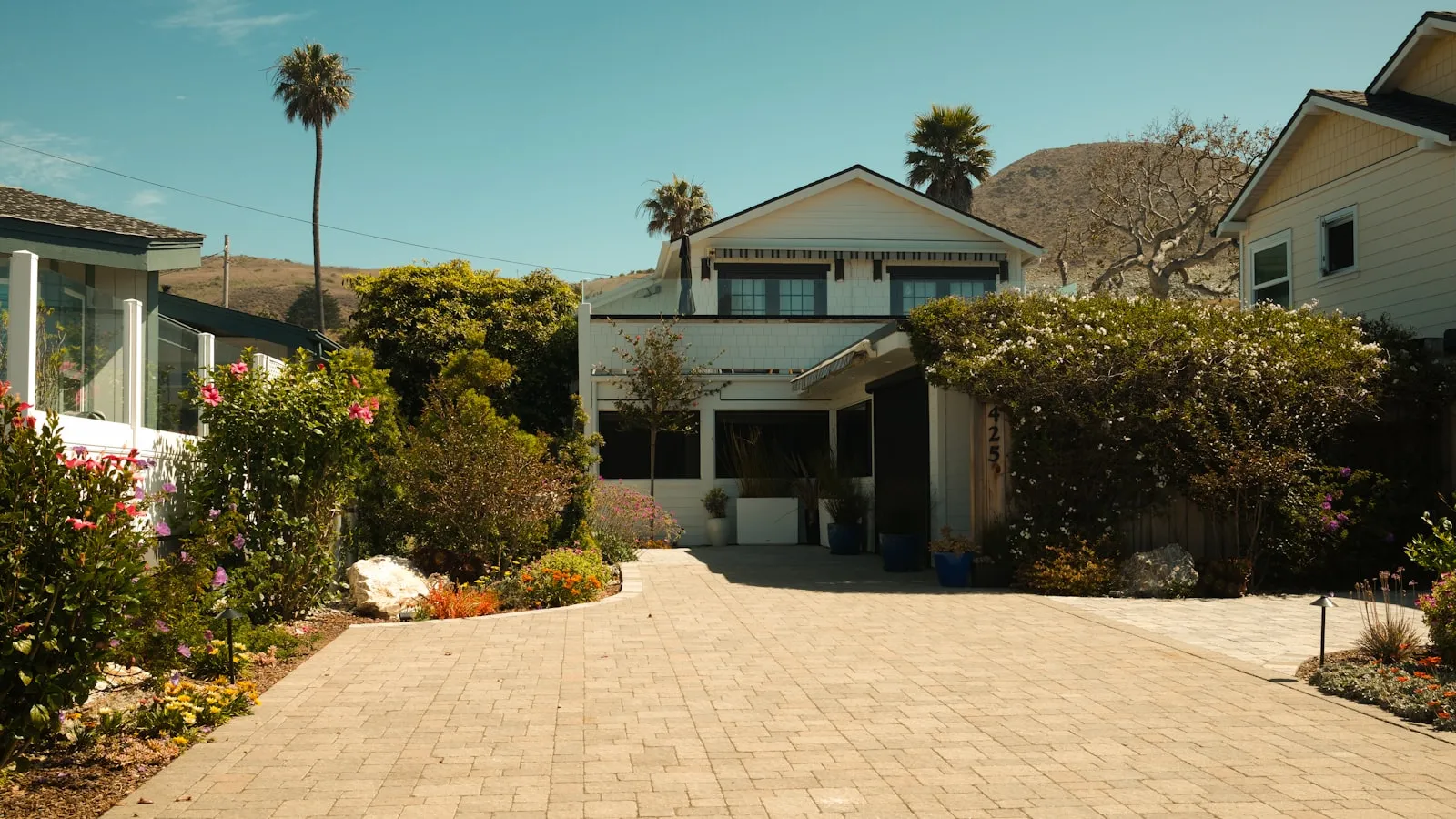 A bungalow-style house with a long paved brick driveway and lush landscaping, palms in the background