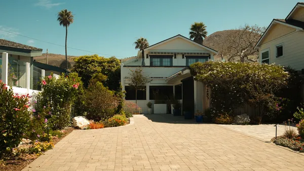A bungalow-style house with a long paved brick driveway and lush landscaping, palms in the background
