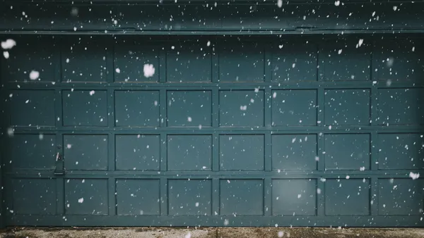 A teal-panelled garage door on a house exterior with snow falling, in low, moody light