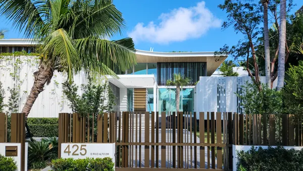 A modern bronze metal slat driveway gate in front of a white contemporary house with palm trees