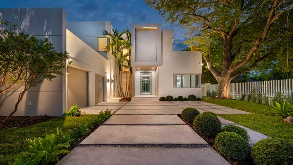 A modern white house glowing at dusk with a long illuminated entry pathway lined with clipped shrubs