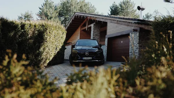 A black luxury SUV parked in a residential driveway beside a stone-and-wood garage, tall hedge alongside