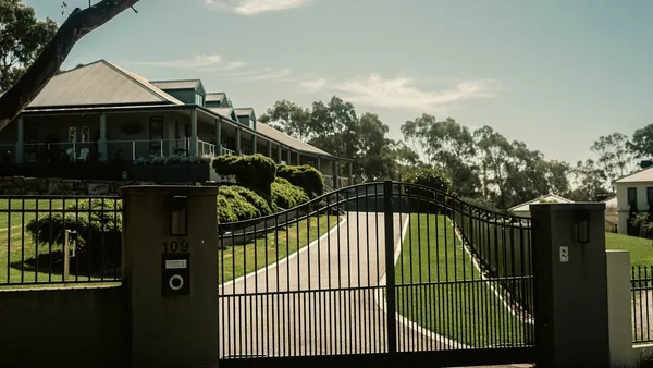 A black metal driveway gate on stone posts opening onto a driveway leading up to a house