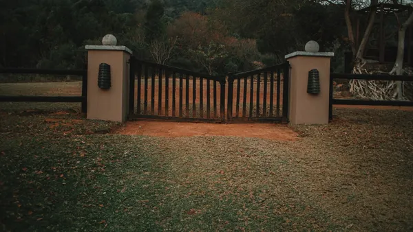 A black metal single-panel swing gate between lit stone pillars at dusk, with a post-and-rail fence on either side
