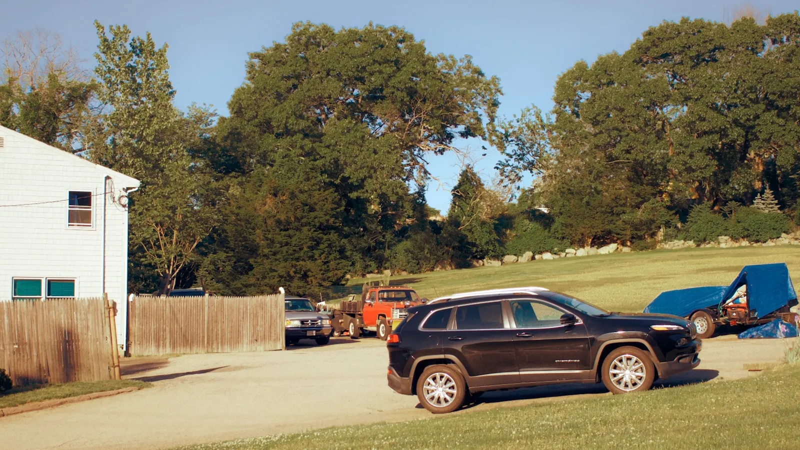 A black SUV parked in a gravel residential driveway beside a white house, a wooden fence and green trees in the background