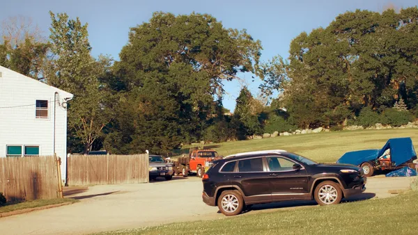 A black SUV parked in a gravel residential driveway beside a white house, a wooden fence and green trees in the background