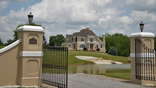 A wrought-iron driveway gate on stone pillars with lamp finials and a long drive toward a large house