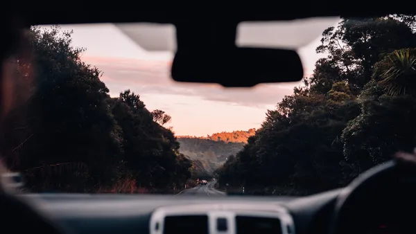 Windshield view of a tree-lined road at dusk, dashboard edge and rear-view mirror visible from the driver's seat