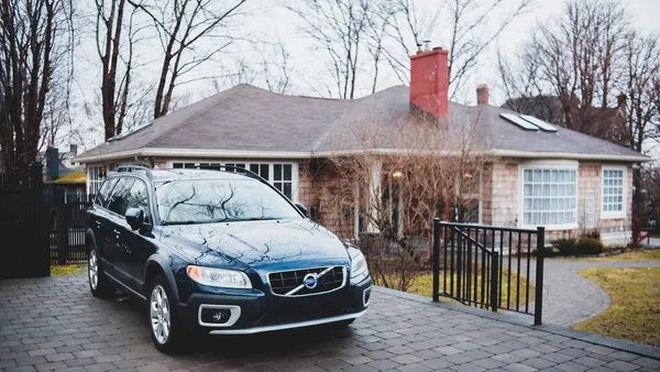 Dark Volvo wagon parked in a paver driveway before a stone house, iron fence and gate visible at left