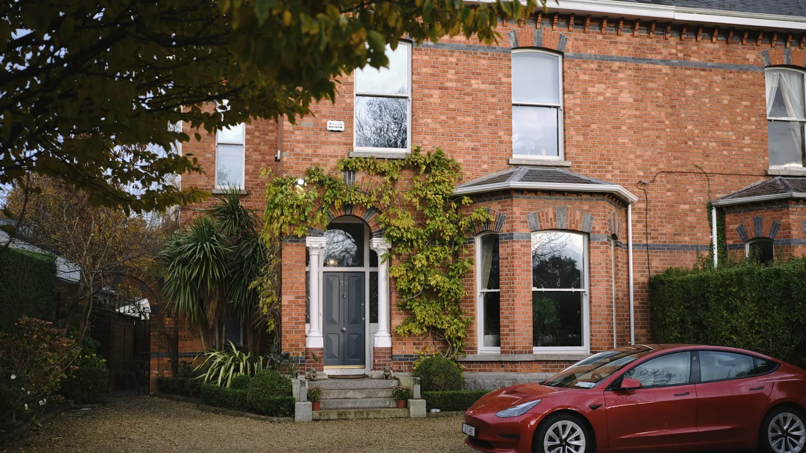 A red Tesla Model 3 parked in front of a brick Victorian house with ivy on the façade