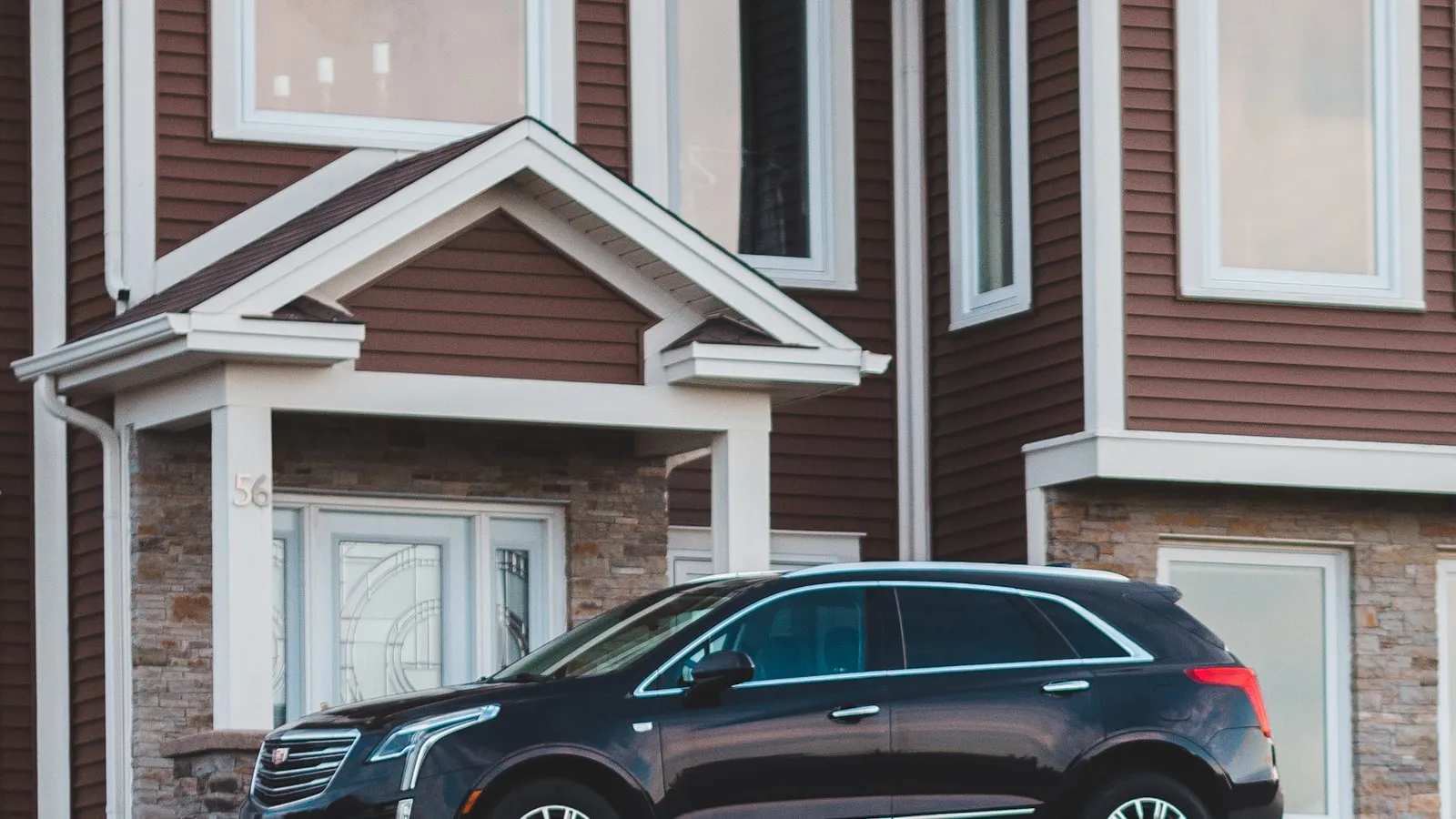A black Cadillac XT5 SUV parked in front of the covered entrance of a brown-sided house