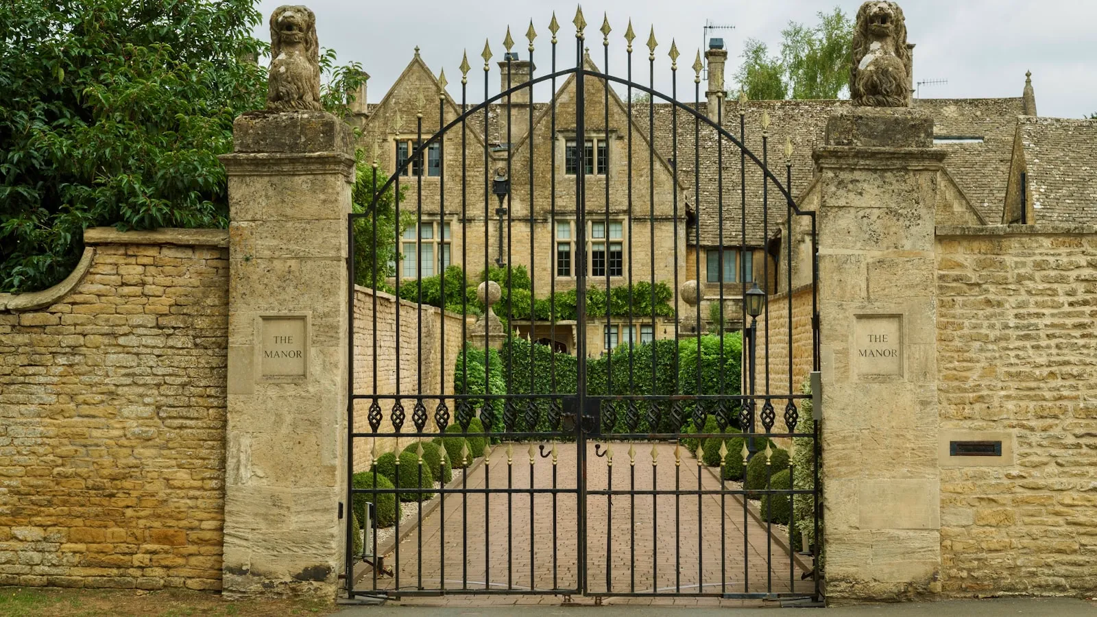 An ornate black wrought-iron gate on red-brick pillars, backed by bare winter trees under an overcast sky